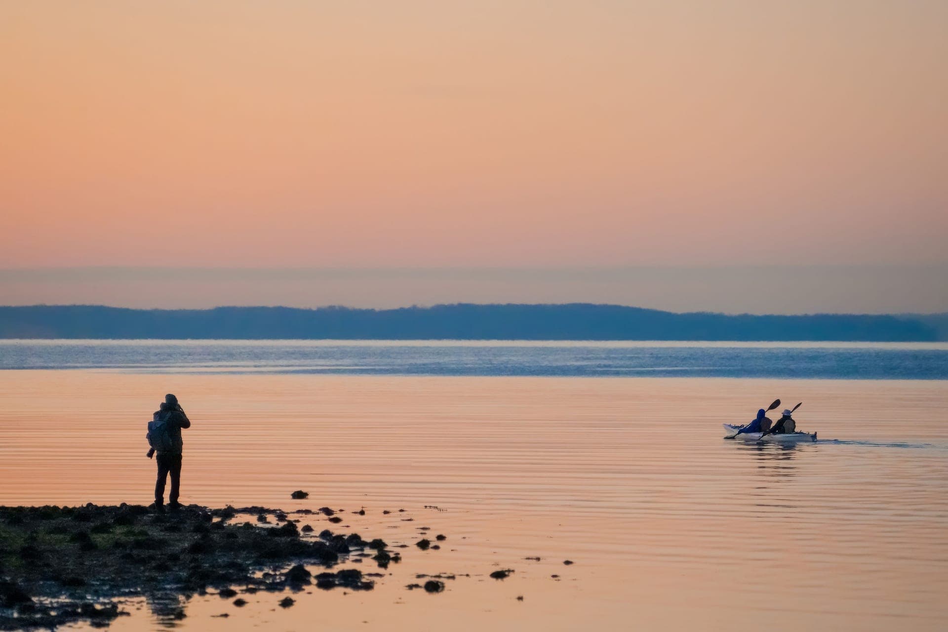 Two kayakers paddling at sunset while a photographer shoots from the shore